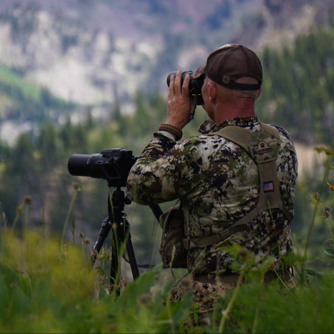 Person in camouflage gear using binoculars with a mountainous landscape in the background
